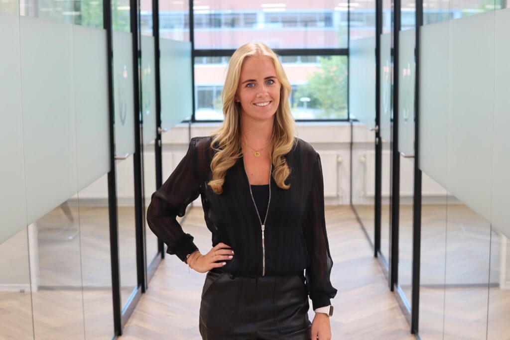 Portret van Demi Lievense: blonde vrouw met een zwarte doorschijnende blouse en leren rok, lachend in een modern kantoor met glazen deuren op de achtergrond.
